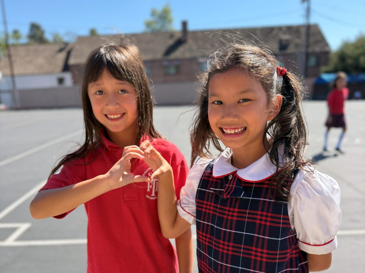 Two smiling SSFP students in uniform making a heart with their hands on the playground