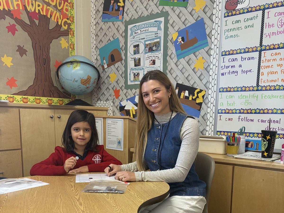 A teacher smiling beside a young student at a classroom table