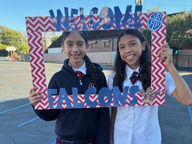 Two smiling SSFP students holding a red and white Welcome Falcons frame on the playground